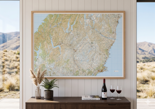 Framed Central Otago map on a wall with a wooden sideboard below displaying a bottle of wine and glasses.