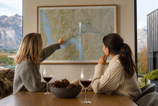 Two women sitting at a table with wine glasses, pointing at a map of Queenstown and Arrowtown on the wall.