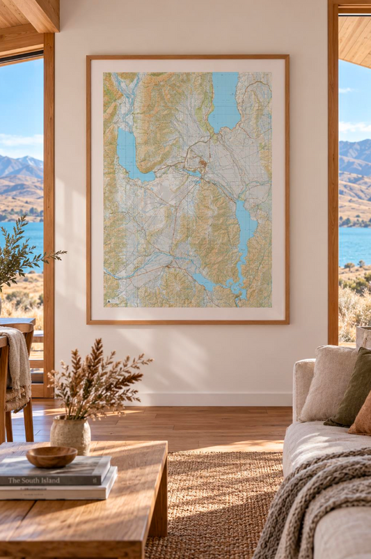 A large framed topographic map print of Twizel, Omarama, Ōhau & Benmore hangs on a beige wall in a modern, sunlit living room with a wooden coffee table, woven rug, and a sofa with a throw blanket.