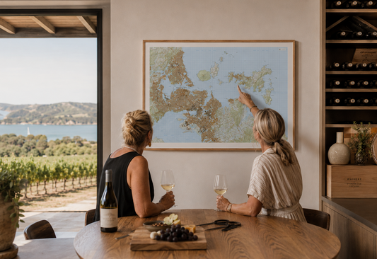 Two women sitting at a table with wine glasses, pointing at a map of Auckland and Waiheke Island, in a room with a view of vineyards.