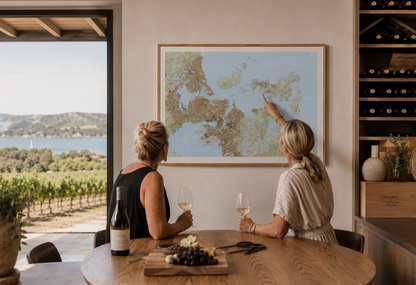 Two women sitting at a table with wine glasses, pointing at a map of Auckland and Waiheke Island, in a room with a view of vineyards.